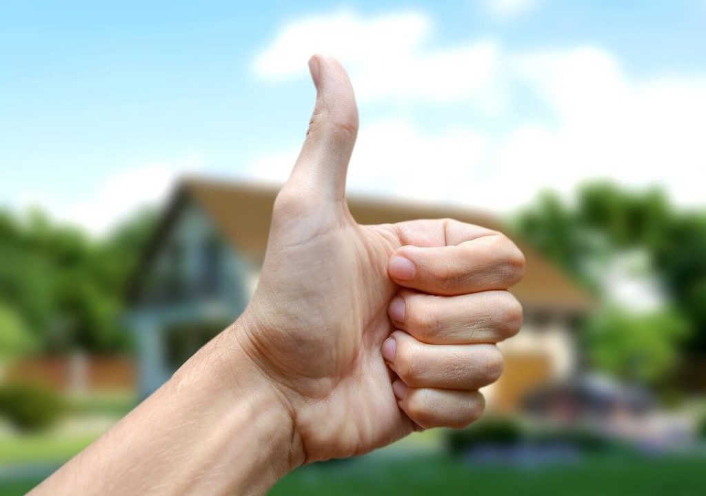 На фото изображена рука с пальцем вверх на фоне дома.